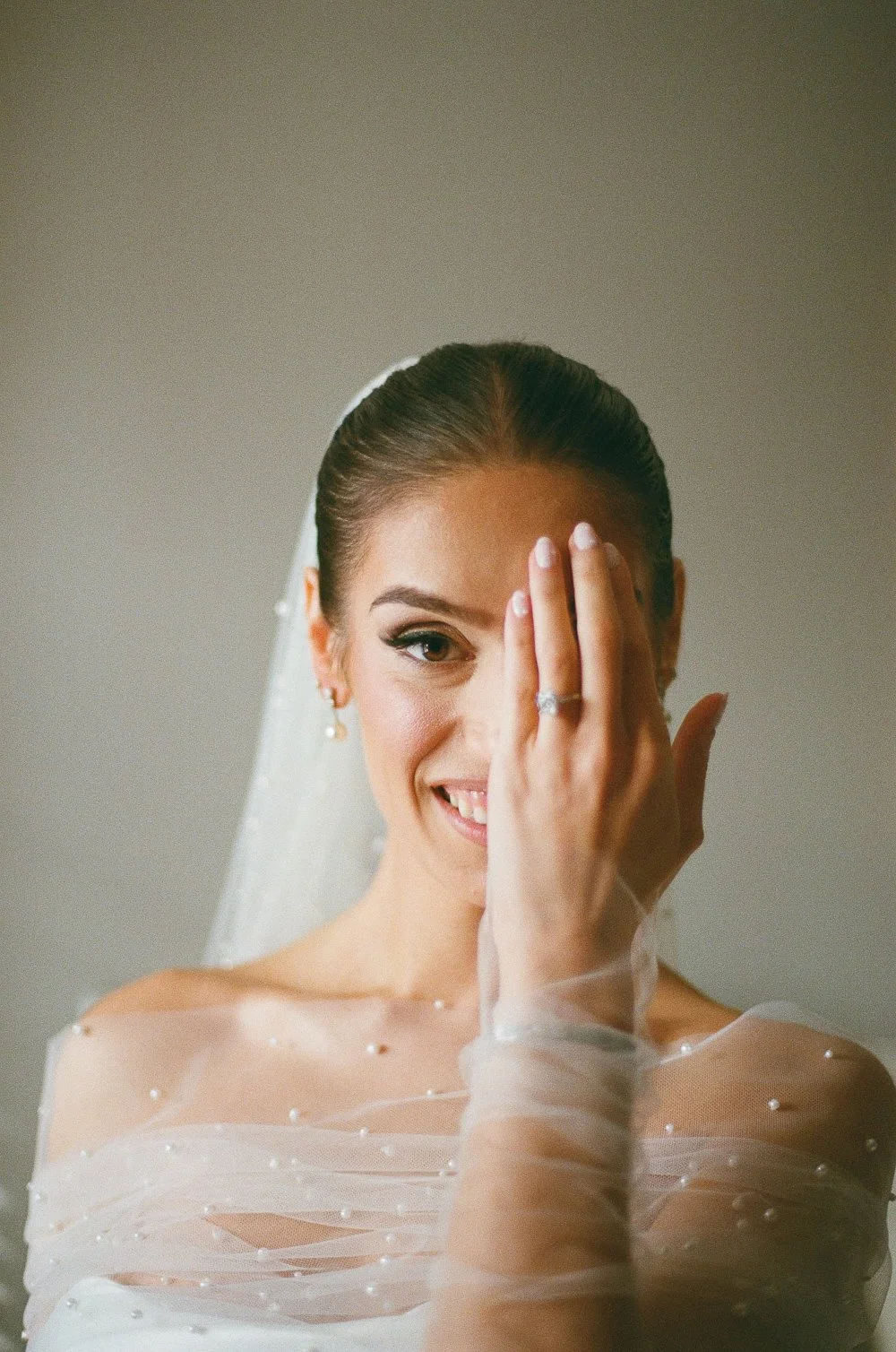 A smiling bride with bridal makeup and veil, covering one eye with her hand, showing an engagement ring, in a wedding dress with pearl embellishments.