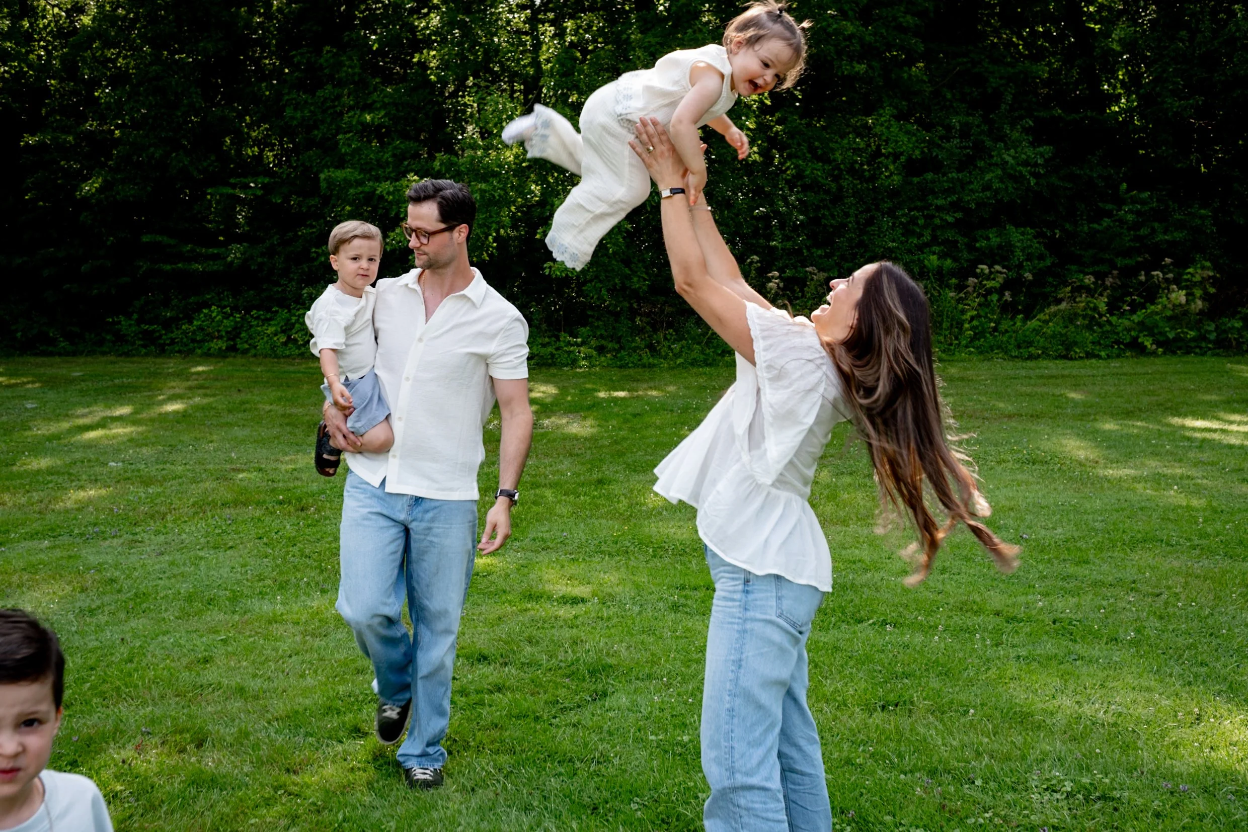 A woman lifts a smiling young girl into the air outdoors, with a man holding a young boy nearby on a grassy area surrounded by trees.