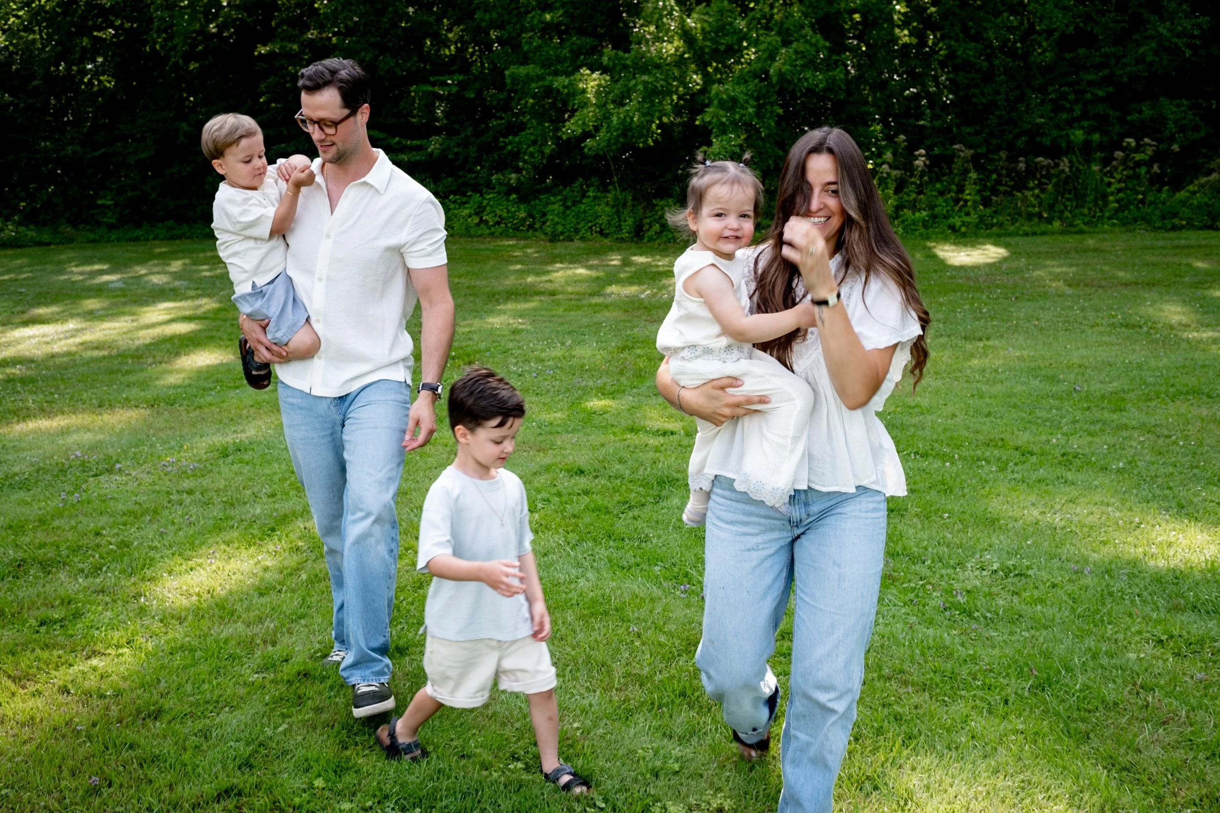 A family of five enjoying a day outdoors in a park with green trees. A man is holding a toddler, a woman is carrying a young girl who is smiling, and two boys are walking on the grass.