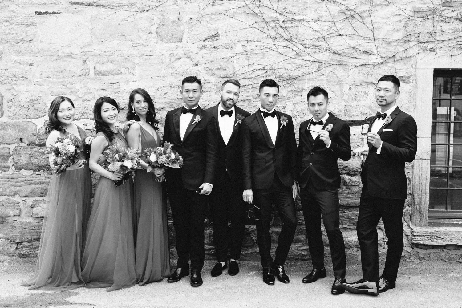 Group of seven people in formal attire standing against a brick wall, with four women holding bouquets of flowers on the left and three men in tuxedos on the right.