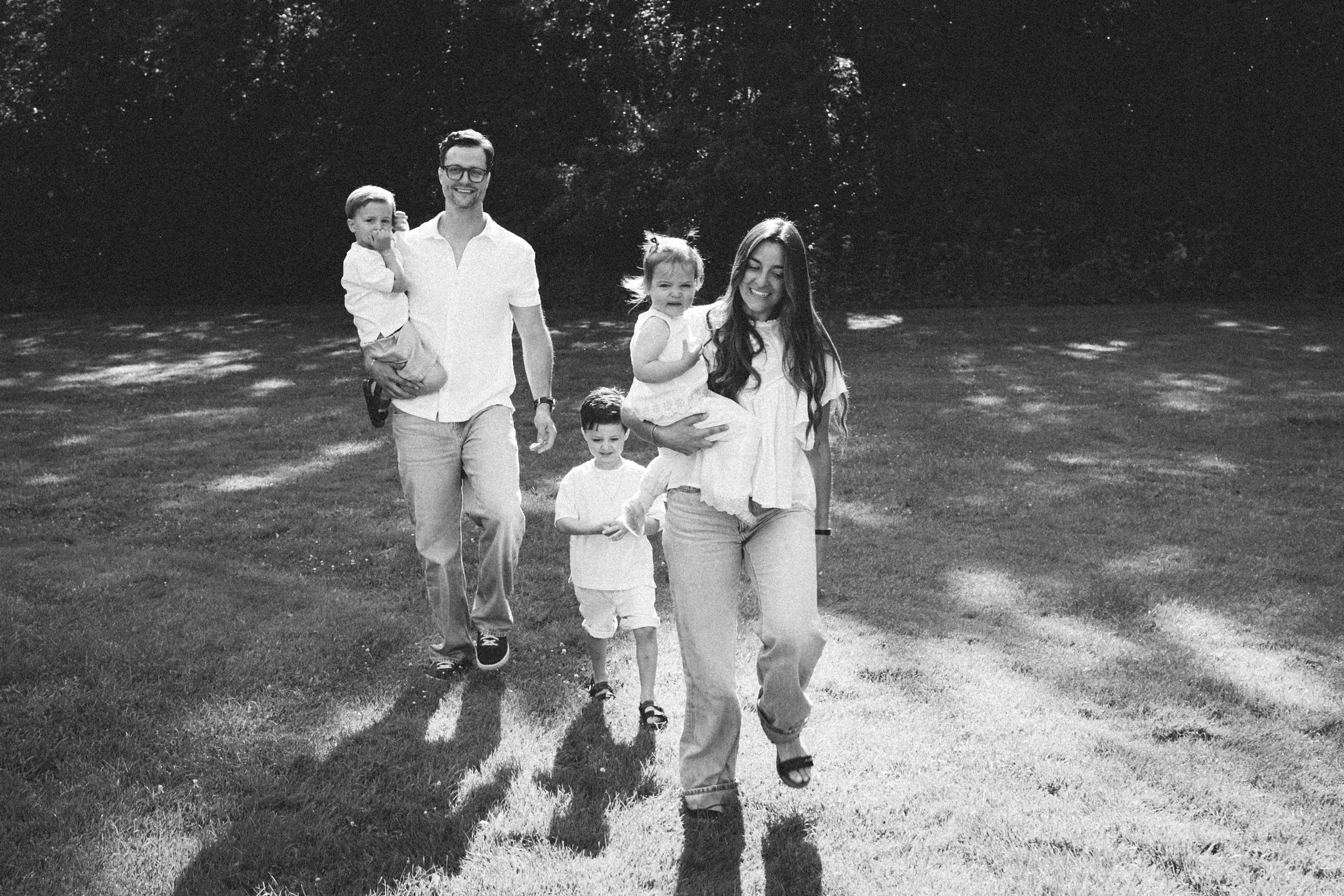 A black and white photo of a family walking outdoors on grass with trees in the background. The family includes a man, a woman, and three children. The man is holding a young girl, and the woman is holding a young girl. A boy is walking in front of t