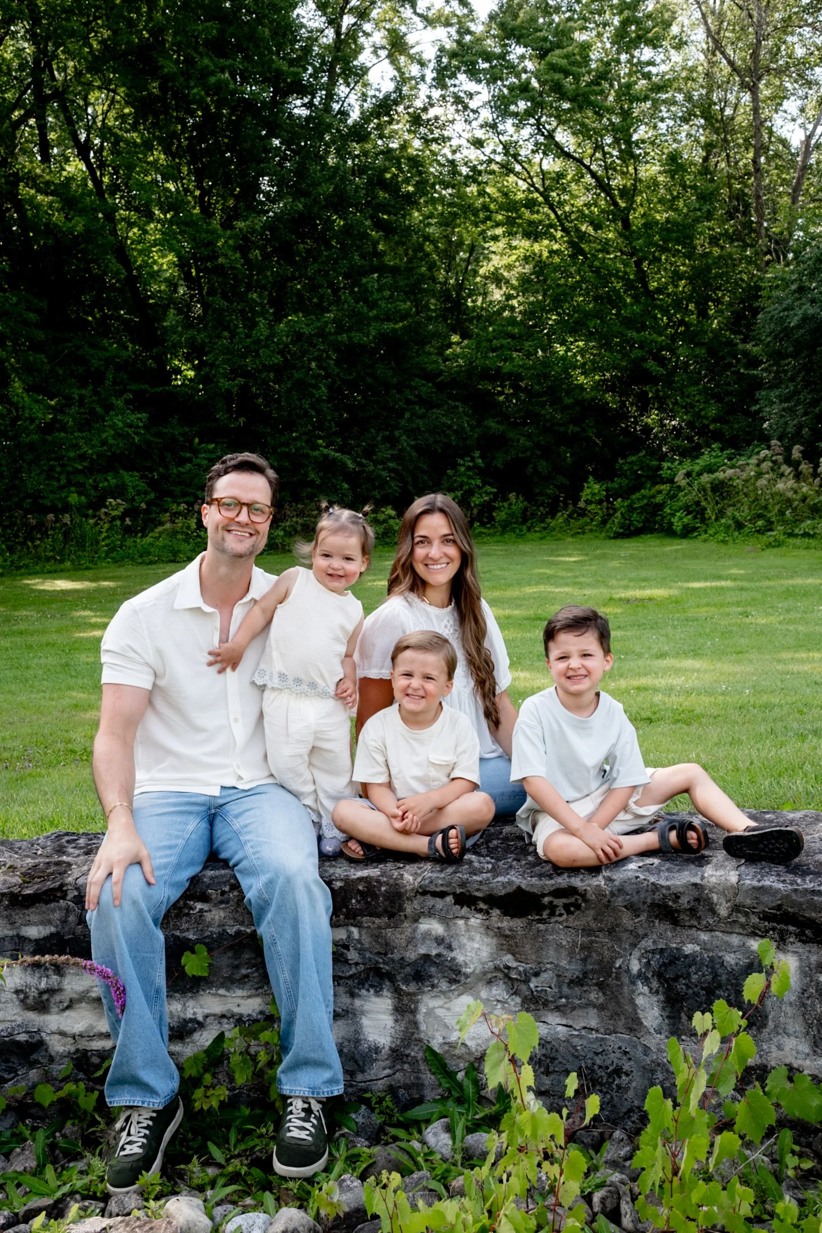 A smiling family of six, including two adults and four children, sitting outdoors on a stone ledge in a park with green trees and grass.