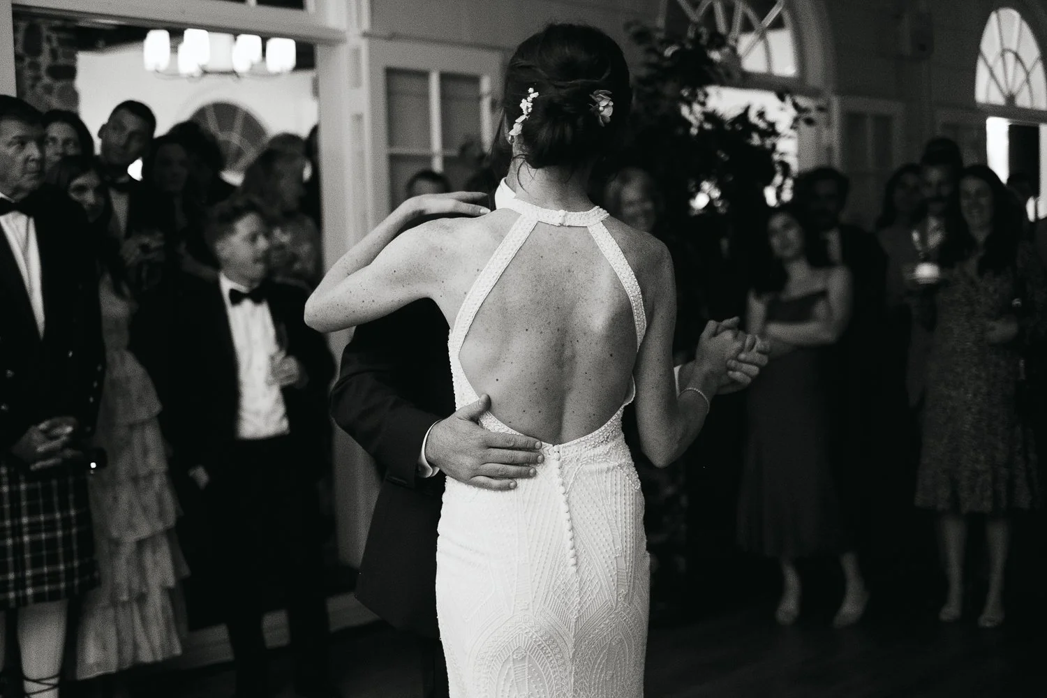 A black-and-white photograph of a bride and groom dancing at their wedding reception surrounded by guests, with the bride wearing a backless wedding dress and floral hair accessories.