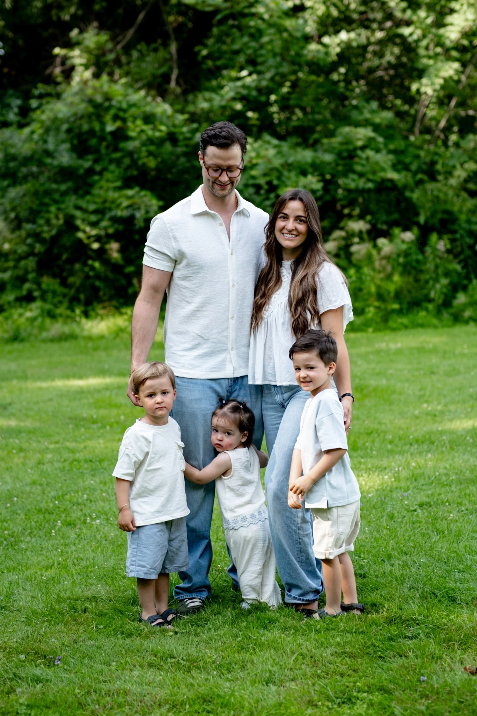 A family of six standing outdoors on a lush green lawn with trees in the background. The family consists of two adults and four children, all smiling and dressed in casual clothing.