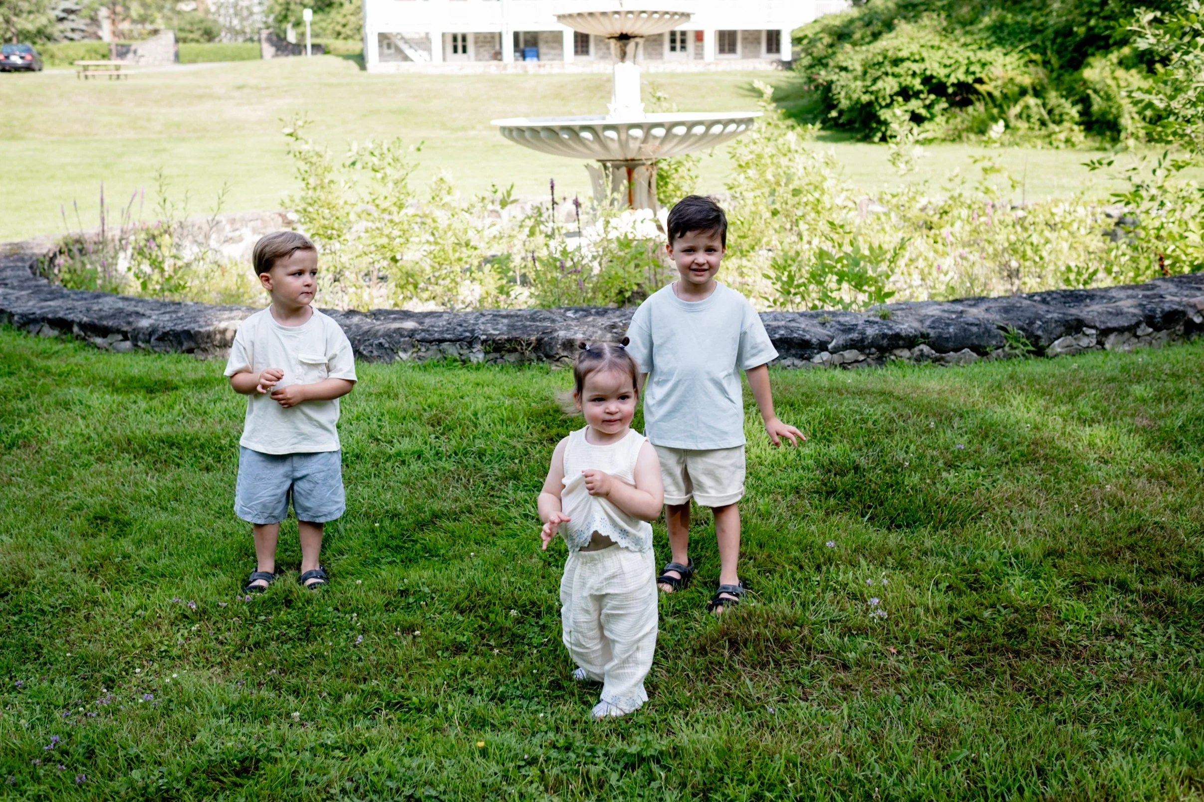 Four young children walking across a lush green lawn near a stone wall with a white fountain in the background, a building with porch and stairs further back, and trees and bushes surrounding the area.