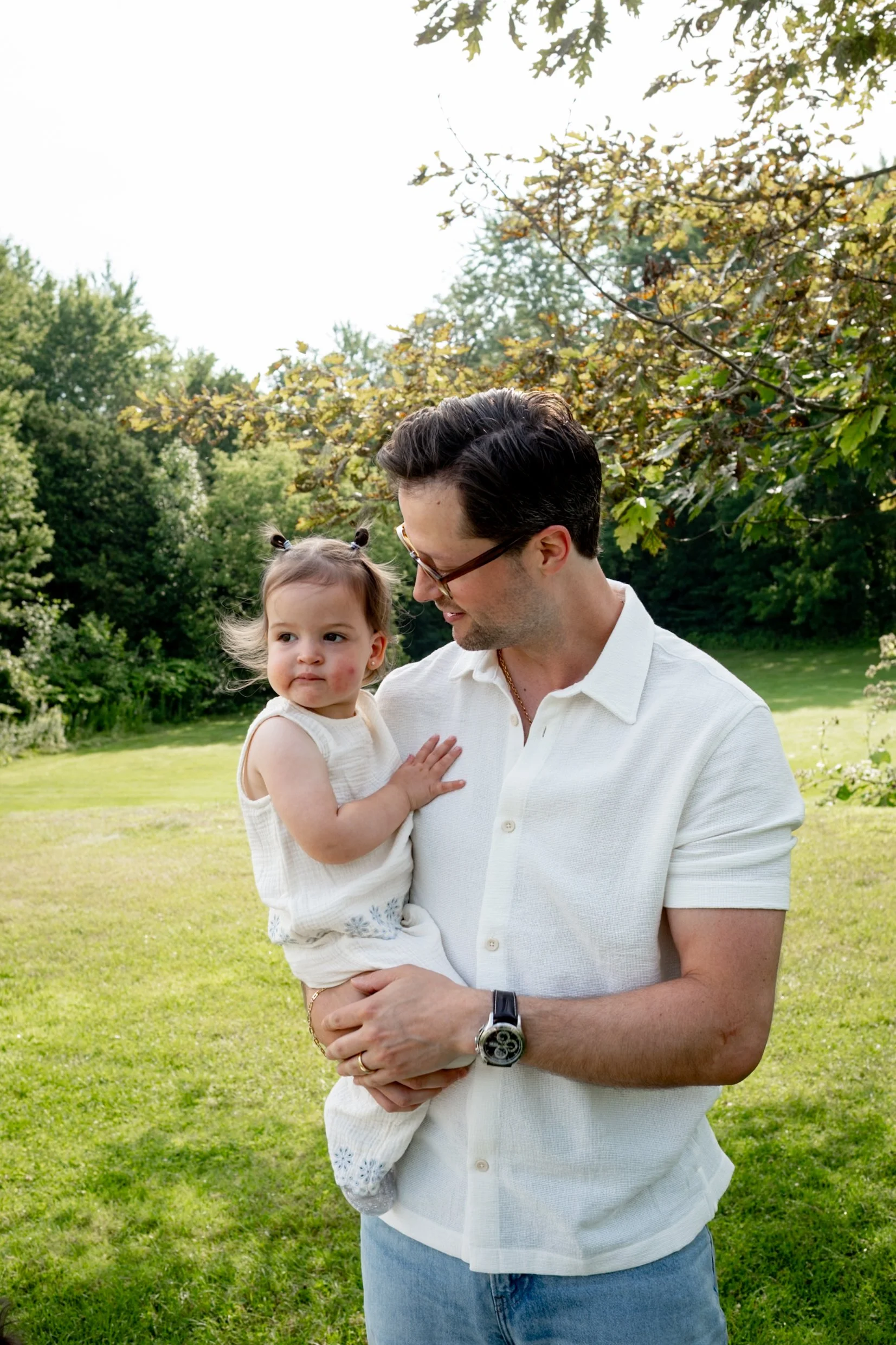 A man holding a young girl outdoors in a green park with trees in the background. The man is wearing glasses, a white shirt, and a watch, and is looking affectionately at the girl. The girl, with pigtails, is wearing a sleeveless dress and gazing off
