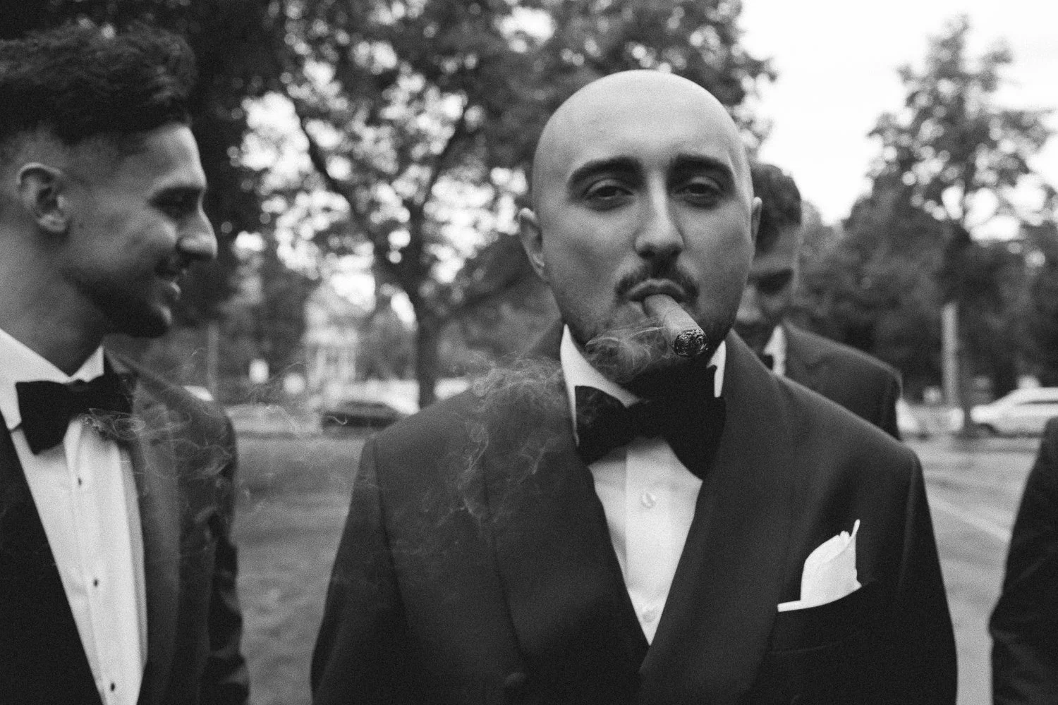 Three men dressed in tuxedos, with the central man smoking a cigar, in an outdoor setting with trees in the background.
