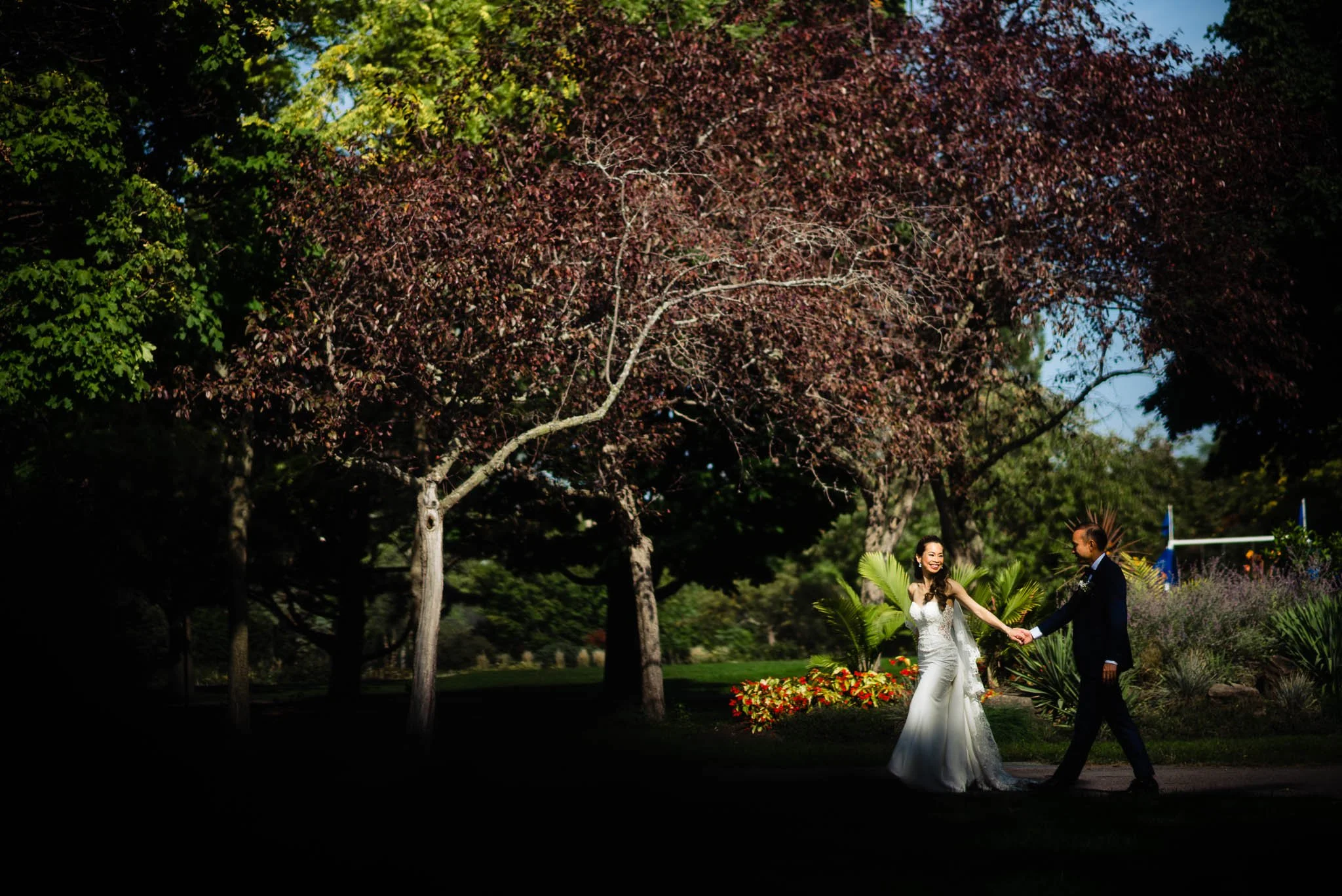 A bride and groom holding hands and smiling in a garden with trees and flowers.