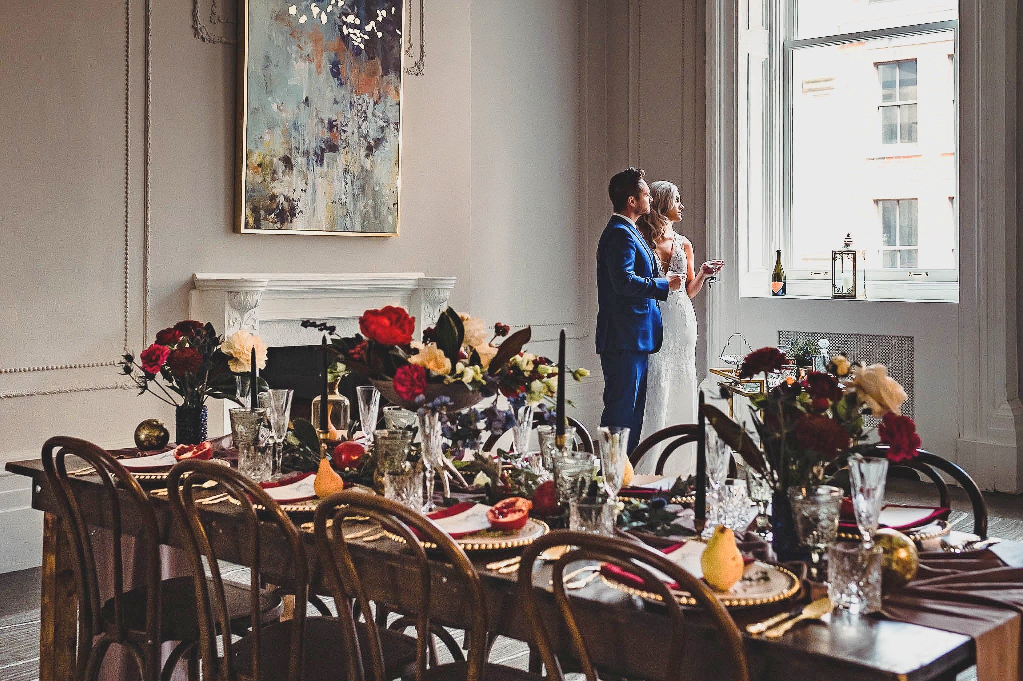 bride and groom take a silent moment alone looking out window. a tablescape is in the foreground 