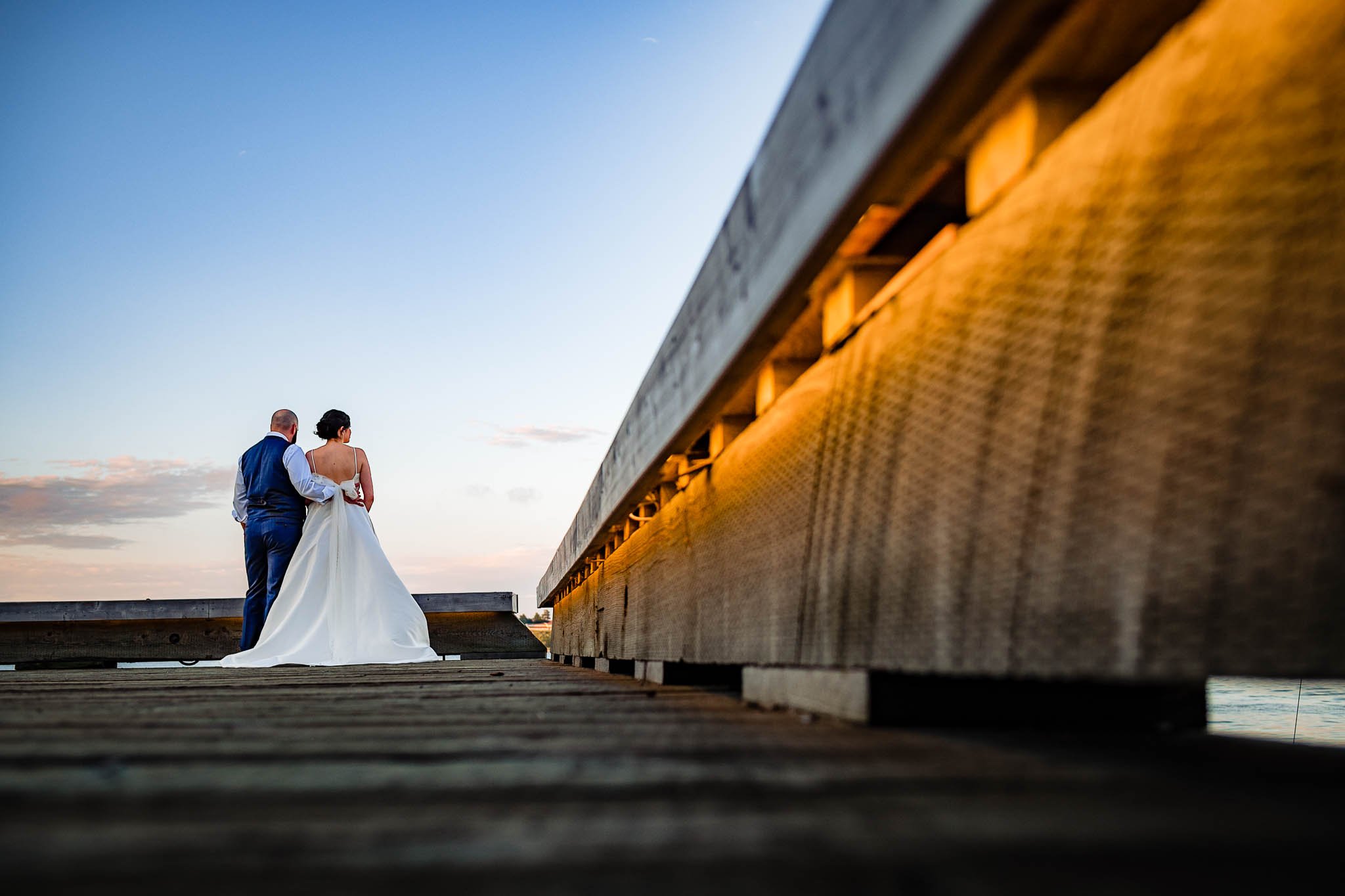 backs are turned away from camera of bride and groom on a dock watching the sun set over halifax 