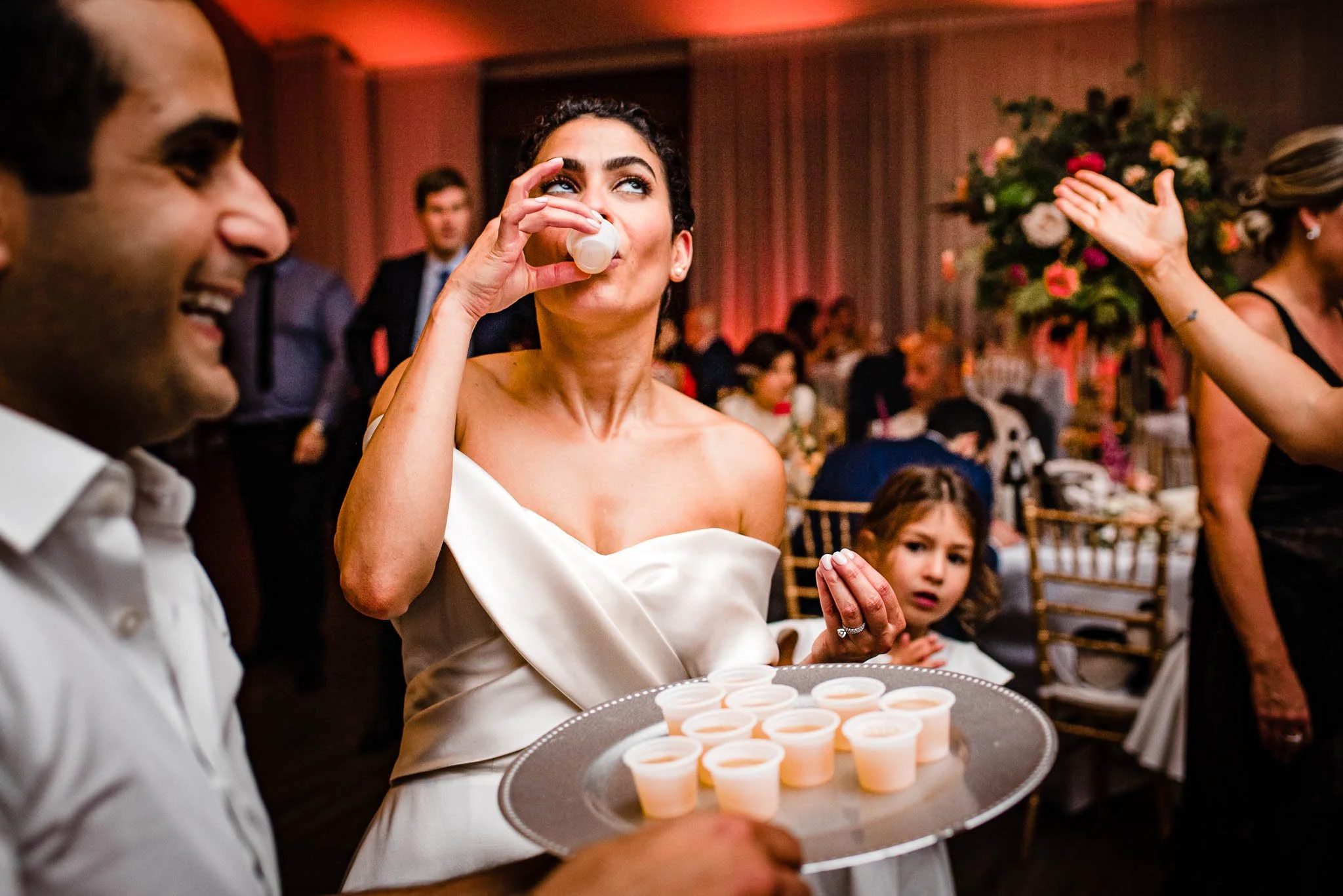 A woman in a white off-shoulder dress is drinking from a small cup at a celebratory event, surrounded by other people, including a smiling man to her left and a young girl in the background. The woman is holding a tray with multiple small cups of a l