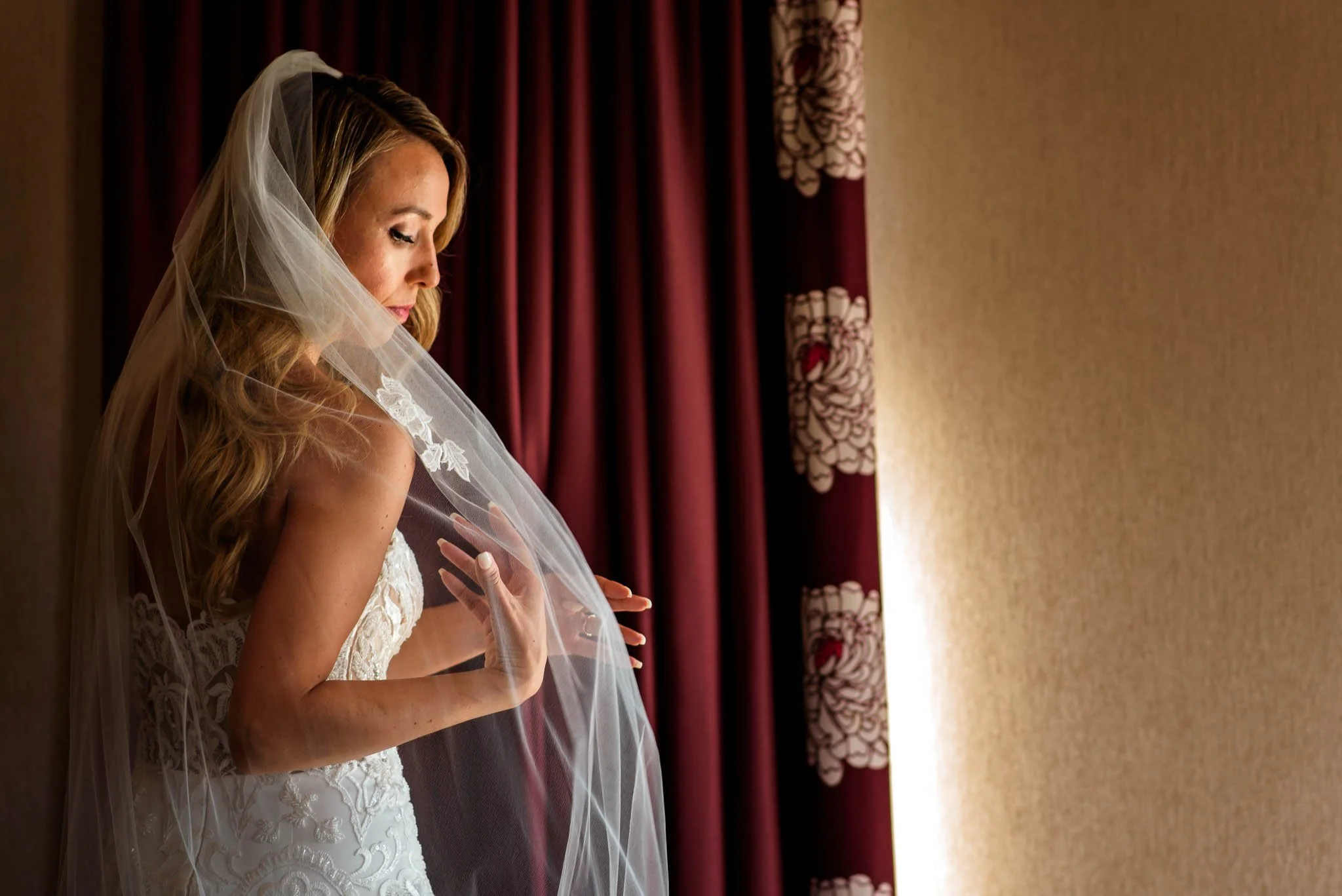A bride with long blonde hair, wearing a white wedding gown and veil, looks down thoughtfully in a room with maroon curtains and beige walls.