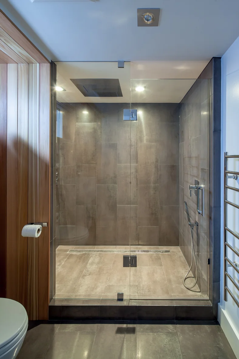 The metallic, oversized, porcelain tile path throughout the basement terminates by wrapping up the walls of the luxury shower, forming a glowing feature wall upon entering the bathroom.