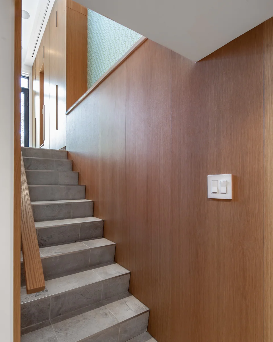 The basement stair is a confluence of materials between two storeys; the tiled floor flows into the staircase itself and wood cabinets seamlessly transition to wood panels that frame the staircase and landing below.