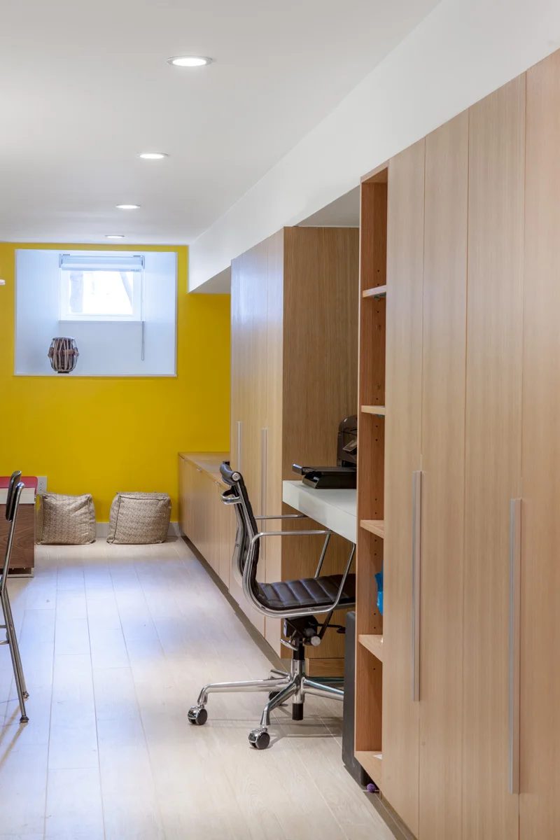 A bank of built-in storage runs the full length of the basement to take advantage of limited height under mechanical ductwork and features open bookshelves, closed storage for supplies and seasonal clothing, a desk, TV, and wardrobe space for guests staying in the basement.