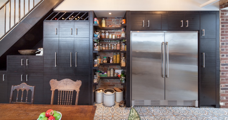 A full wall of built-in storage and pantry space under the stair incorporates the fridge, freezer, a custom wine storage piece, and open pantry shelves for convenient storage of spices, dry goods, and a nook to store pet food.