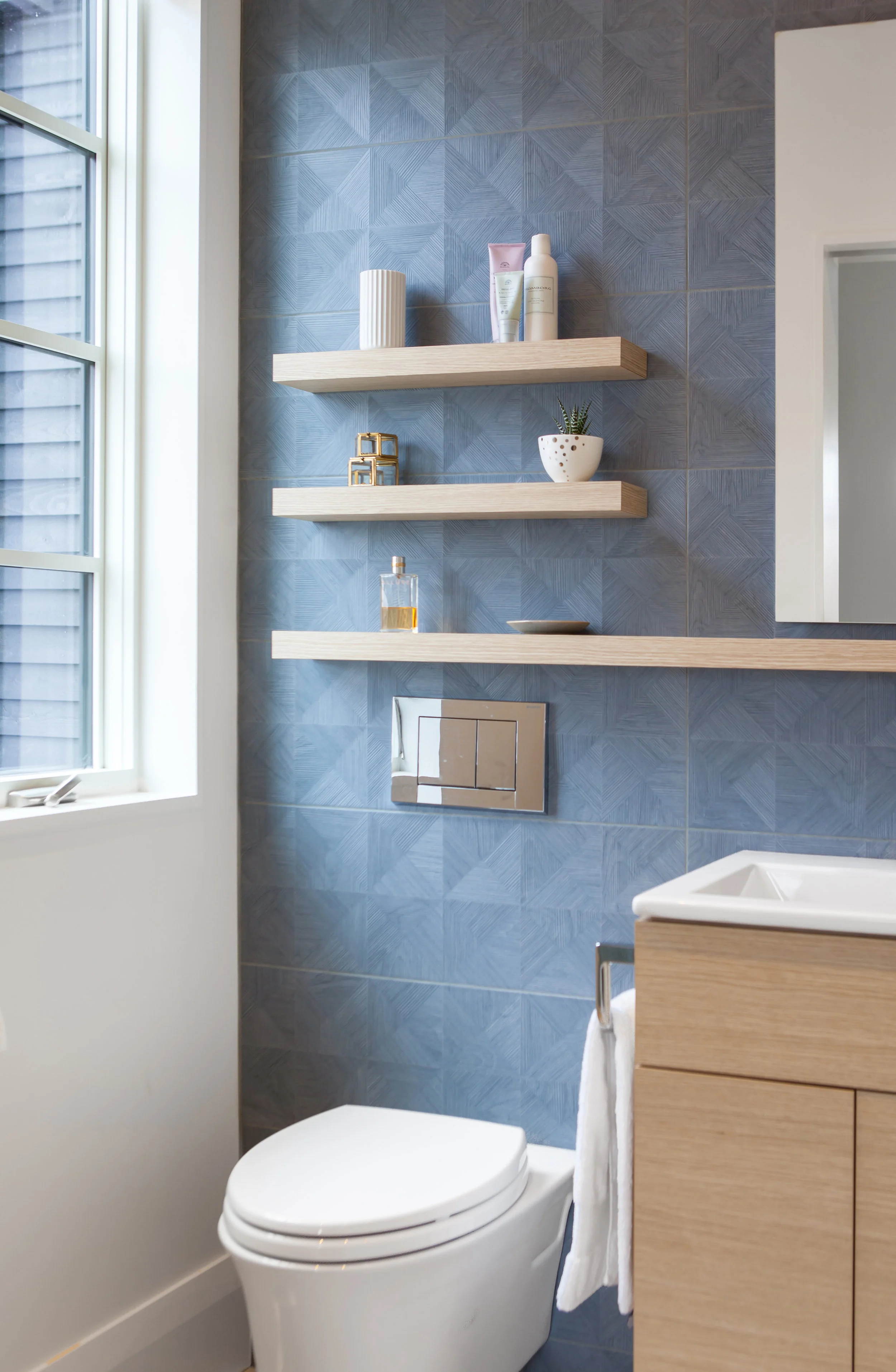 A wall-mounted toilet is another space-saving feature that allows for the full 3-piece bathroom to fit within a modest footprint. A large widow brings generous amounts of light into the bathroom that washes across the blue tile, accentuating its subtle geometry.