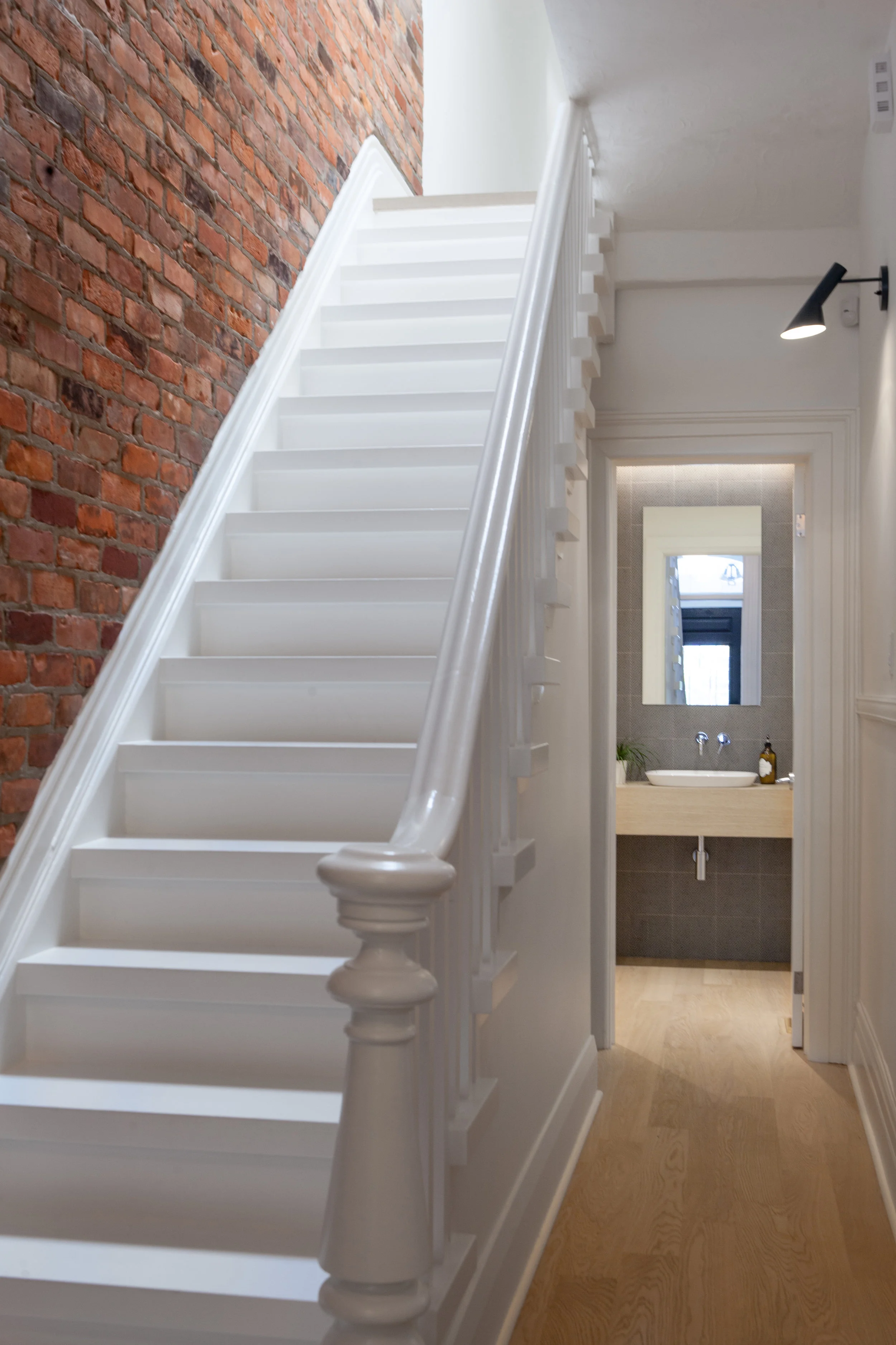 The main hallway features an original brick wall and, like the rest of the front of the house, preserves the traditional baseboards and casework details, while providing a glimpse into the new powder room that is tucked just behind the stair. The powder room’s clean palette of blues and whites hints at the modern addition, a sneak peek for visitors stepping into the home.