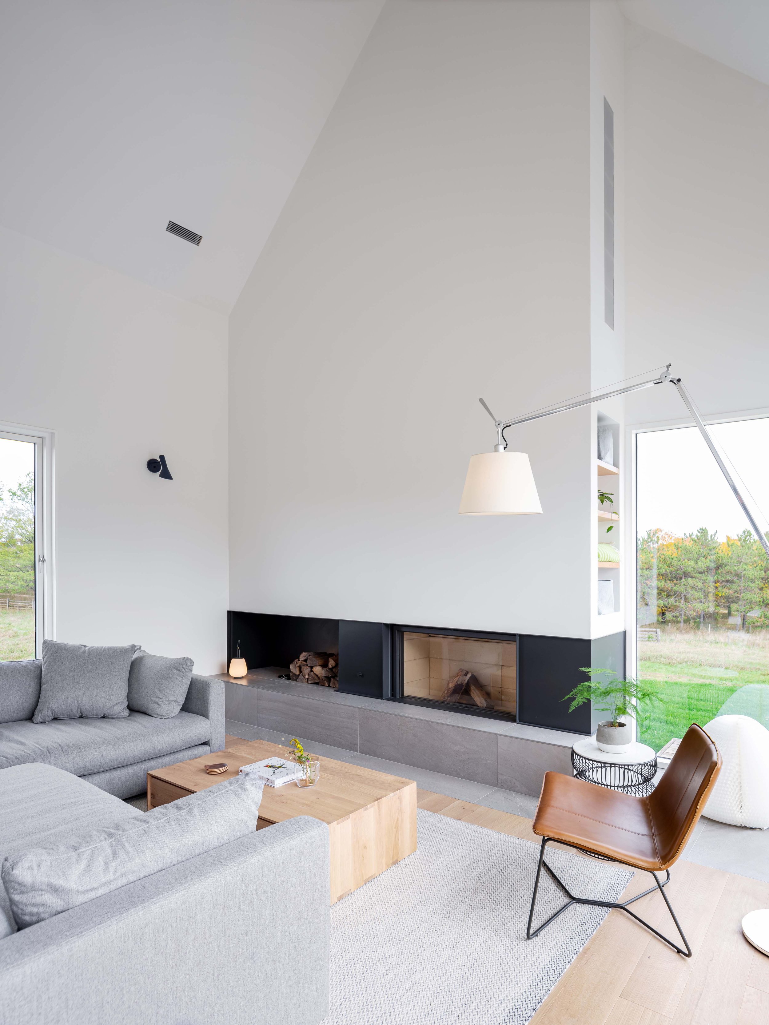 The living room is centred around a linear wood burning fireplace raised on a stone plinth. Both the fireplace and wood storage are cleanly finished in powder coated metal to create a crisp band that wraps across the base of the chimney volume, and