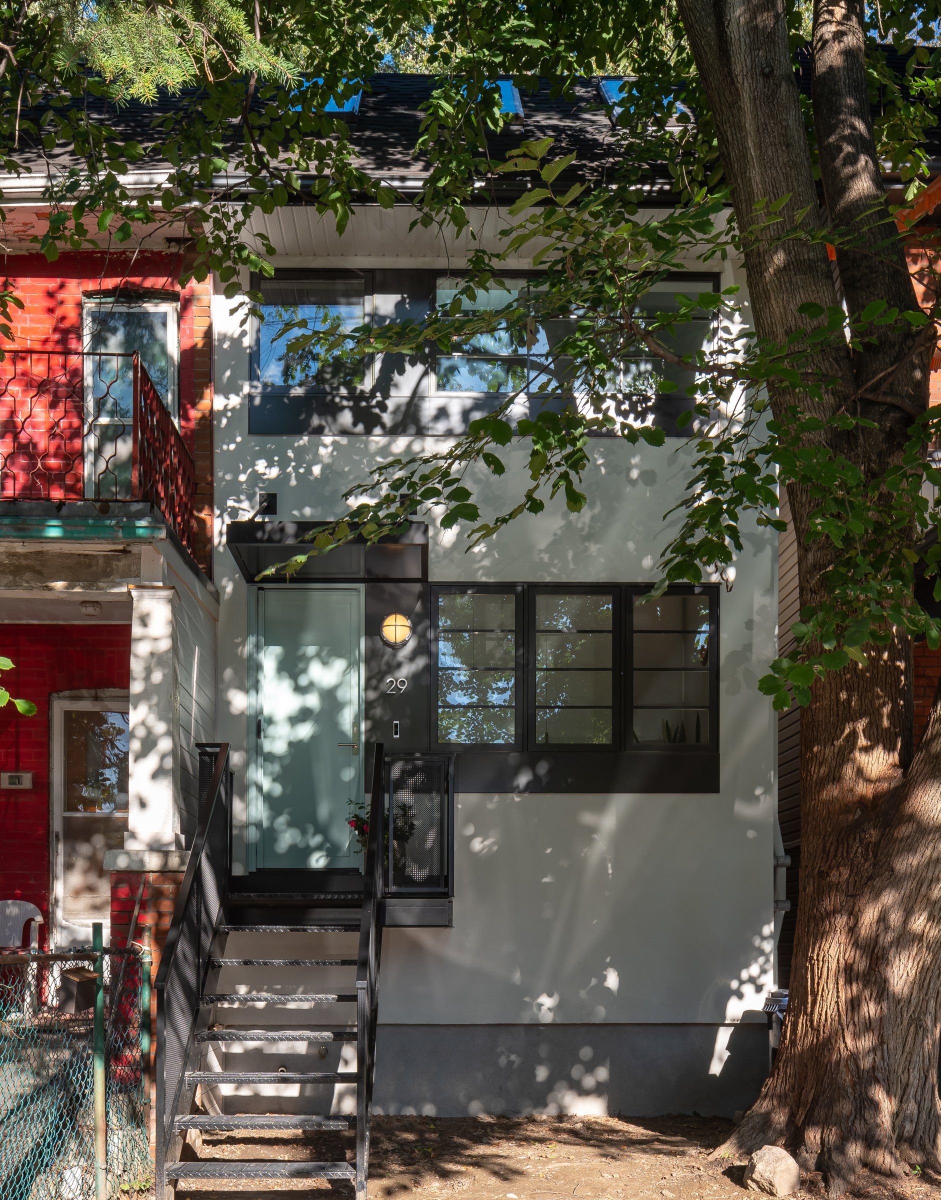The new white stucco exterior stands out from its connected neighbour, and provides the perfect backdrop for shadows cast by the tree in front. While the original roofline is maintained, comparing the front porches of the two homes hints at the spat