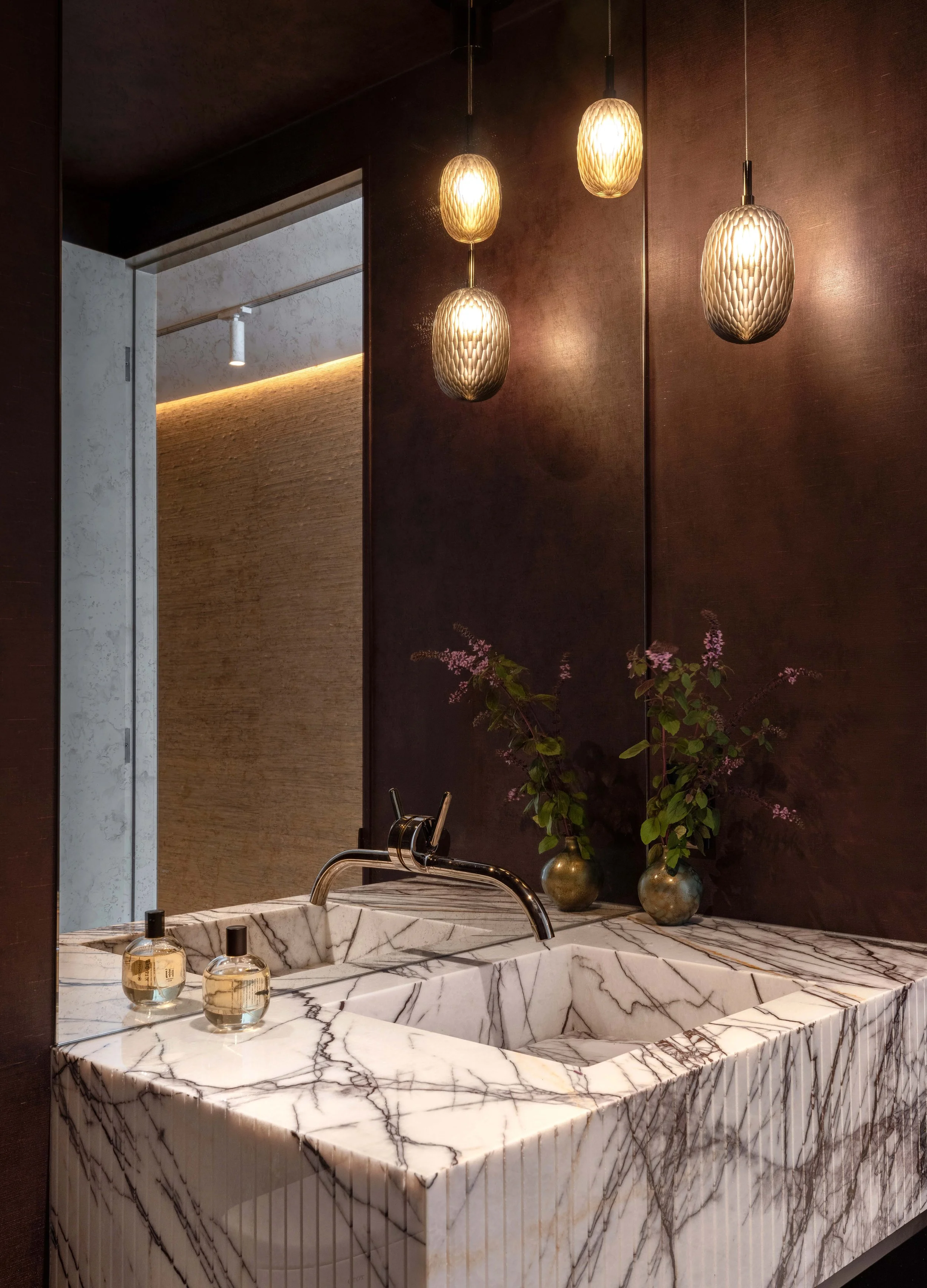  The powder room is dark, moody, and enveloped floor to ceiling in a rich purple wallpaper. Antiqued mirrors and a custom marble vanity with integrated marble sink add further texture and drama. 