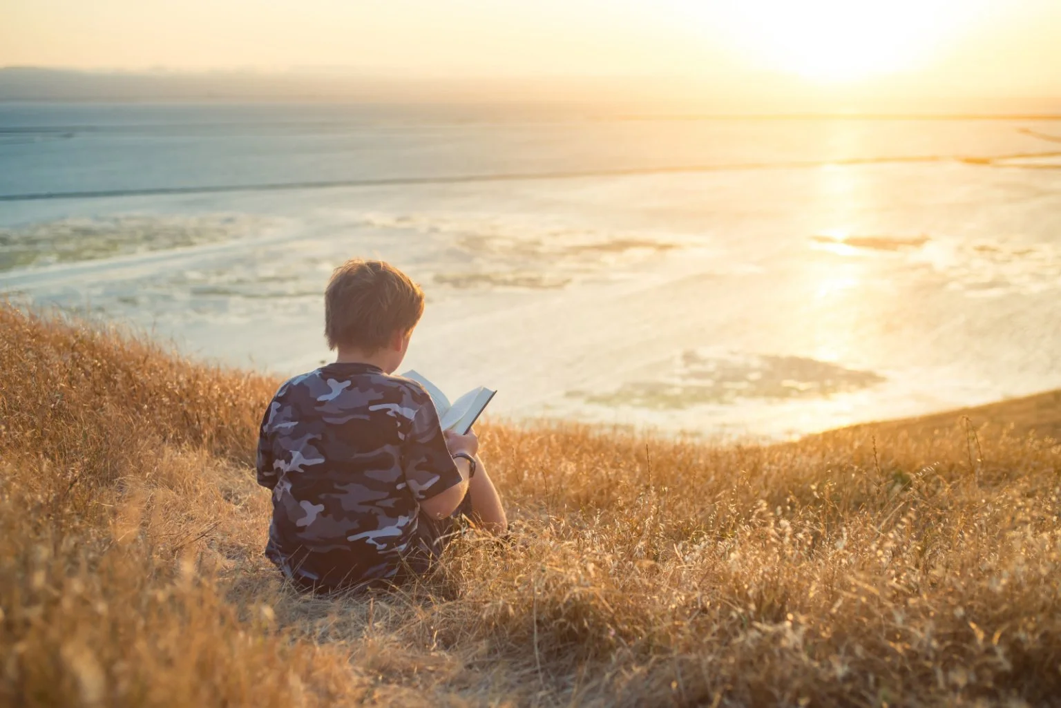A child reading on a hill overlooking the ocean with a beautiful sunset.
