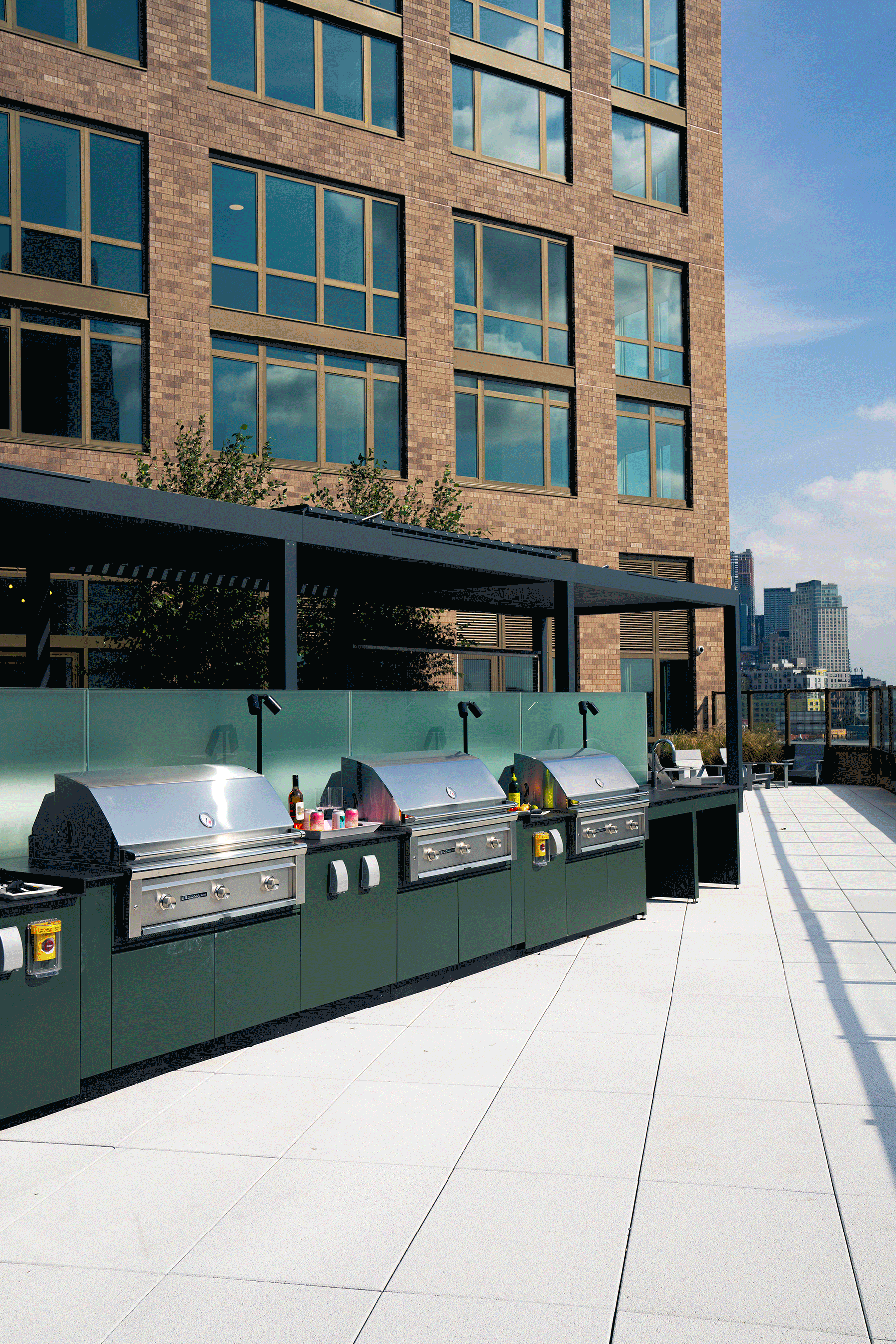 An outdoor rooftop area with four stainless steel gas grills, some condiments and utensils, on a white tiled surface with a multi-story brick building and a city skyline in the background. Creative Direction by Tiffany J. Davis for Context + Co.