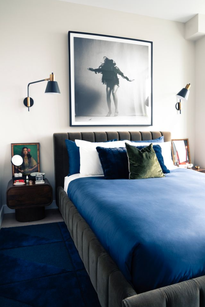 A bedroom with a large bed featuring a gray headboard, blue bedding, and multiple pillows in white, navy blue, and green. Above the bed hangs a large black and white photograph of a woman dancing. On each side of the bed are black wall-mounted lamps, and next to the bed are framed photographs and a small mirror on the nightstands.
