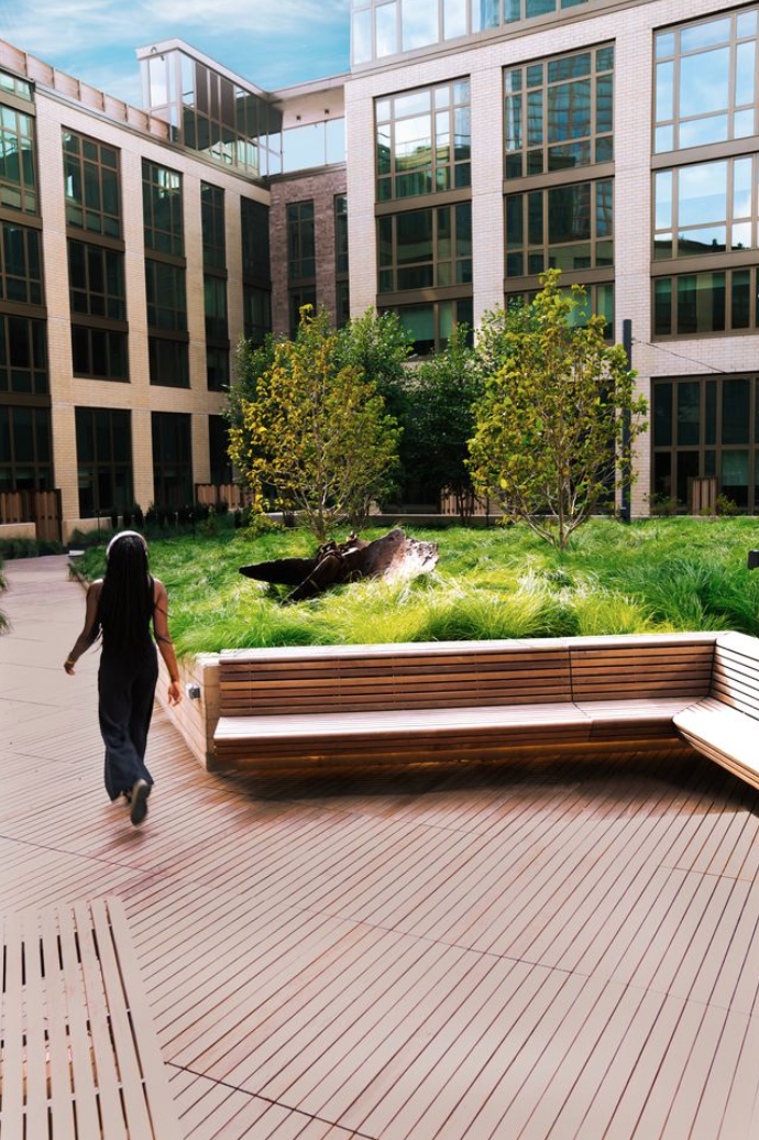 A woman walking on a wooden deck in a modern urban courtyard with greenery and trees, surrounded by tall glass office buildings. Creative Direction by Tiffany J. Davis for Context + Co.