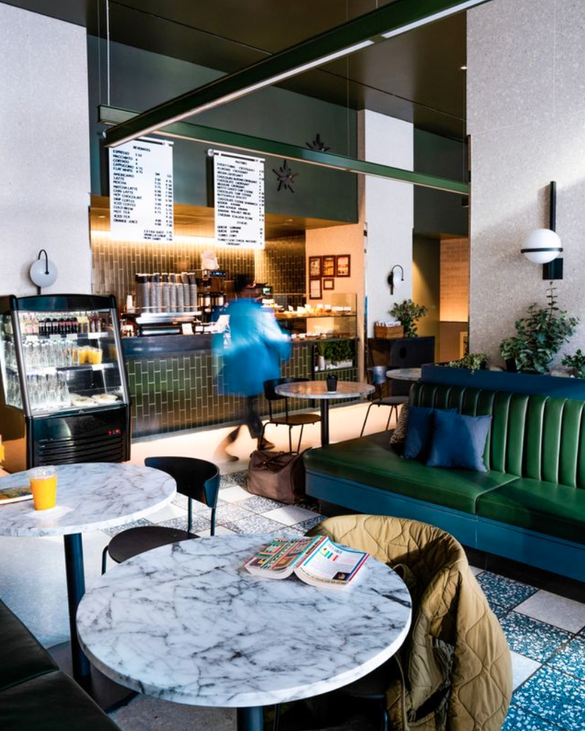 Interior of a modern coffee shop with marble tables, a green sofa with pillows, and a counter with a menu board in the background. There are plants and modern decor. Creative Direction by Tiffany J. Davis for Context + Co.