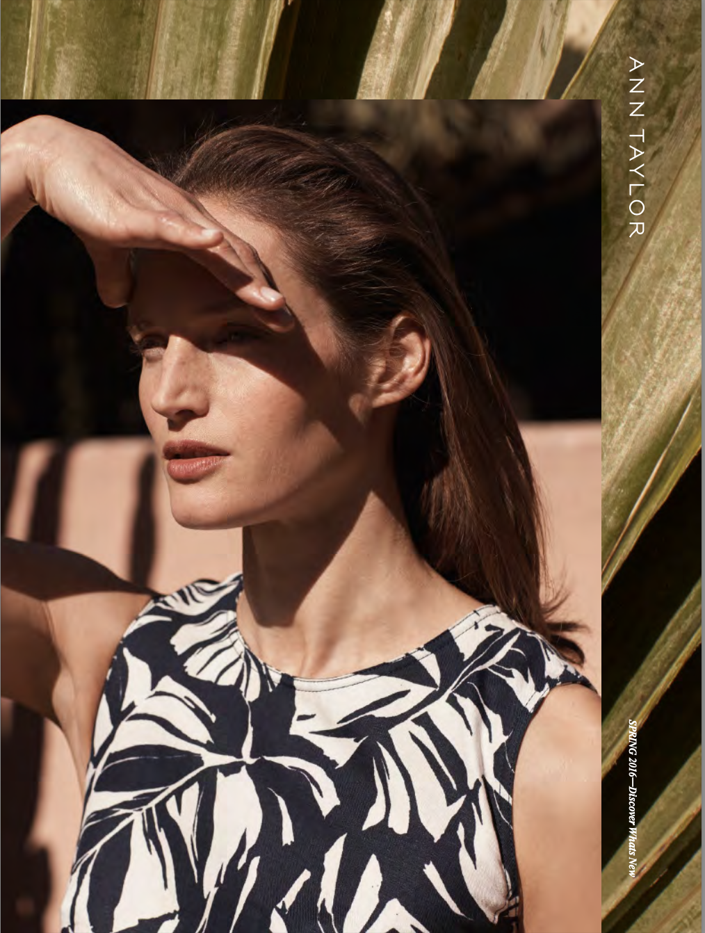 A woman with brown hair in a sleeveless tropical print top, shielding her eyes from the sun with her hand amid an outdoor setting with greenery.