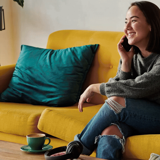 A young woman sitting on a yellow couch, talking on the phone and smiling, with a teal pillow, a coffee cup, and headphones nearby. Sample of a social media template for Canada Life.