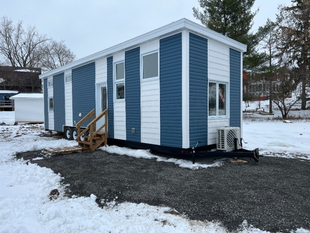 A blue and white modern tiny house on wheels, with stairs leading to the door, set on a gravel foundation in a snowy yard with trees and neighboring houses in the background.