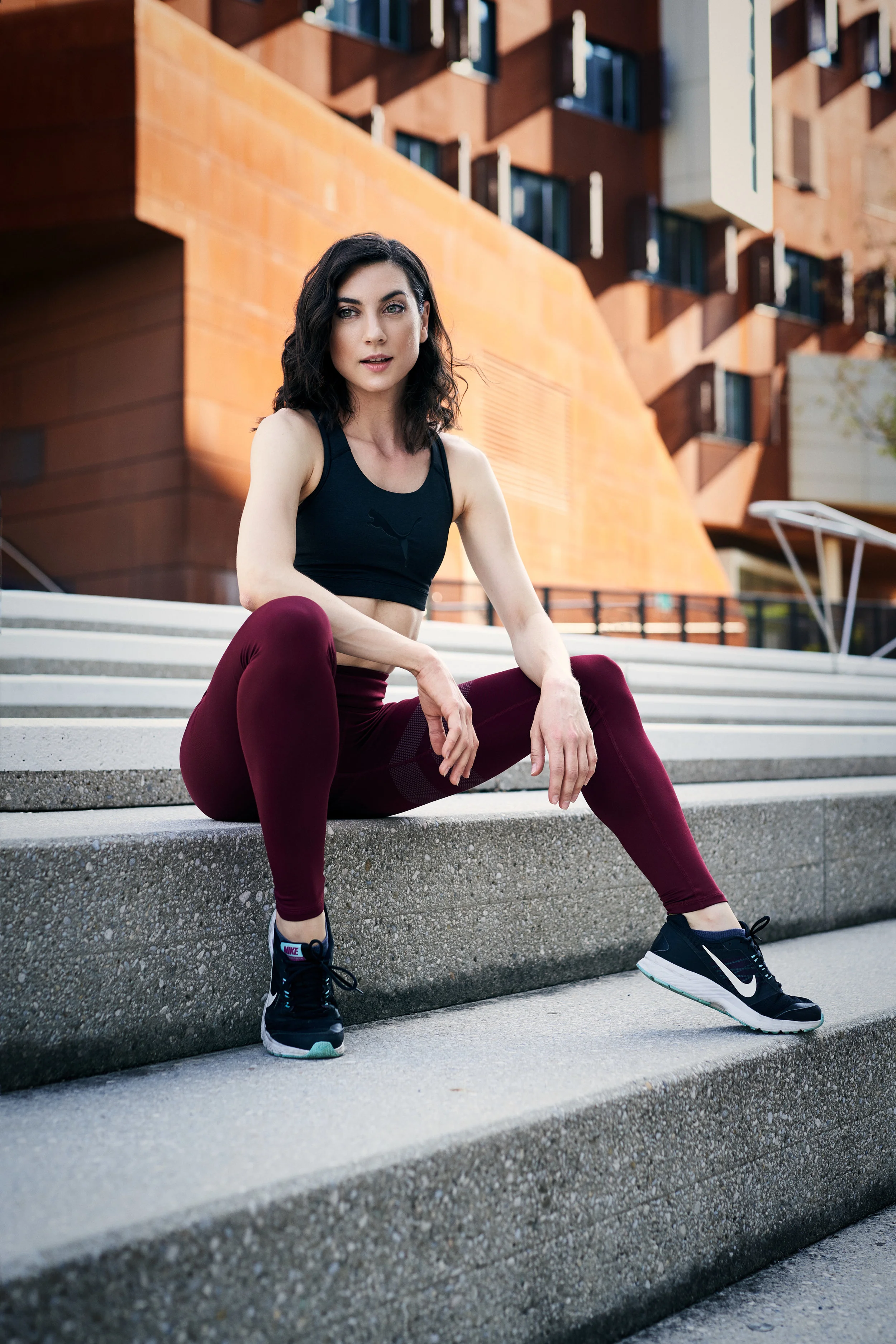 Eine junge Frau mit schwarzen, lockigen Haaren sitzt auf Treppen im Freien, trägt eine schwarze Sporttop, dunkelrote Leggings und schwarze Sportschuhe, vor einem modernen Gebäude mit rotem Backstein-Fassad.