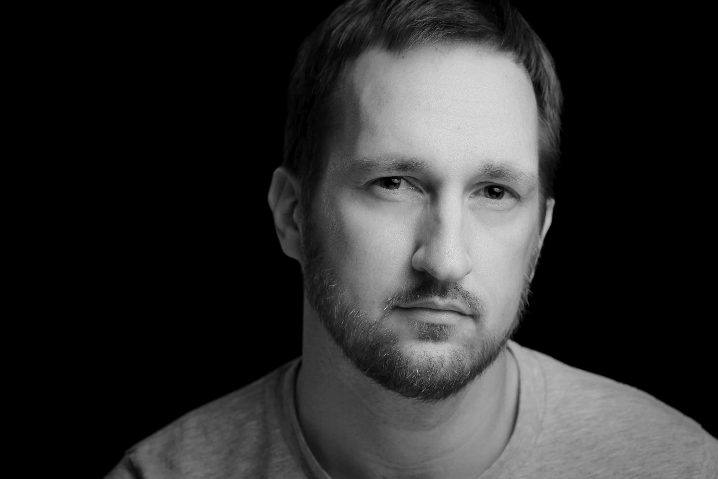 Schwarz-weiß-Porträt eines jungen Mannes mit Bart, kurze Haare und grauem T-Shirt, Blick leicht nach rechts, auf dunklem Hintergrund.