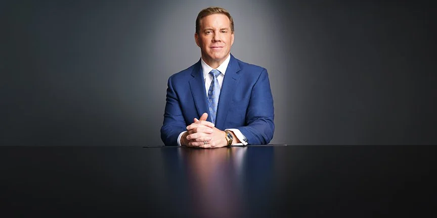 Professional man in a blue suit seated at a dark conference table against a gray studio background.