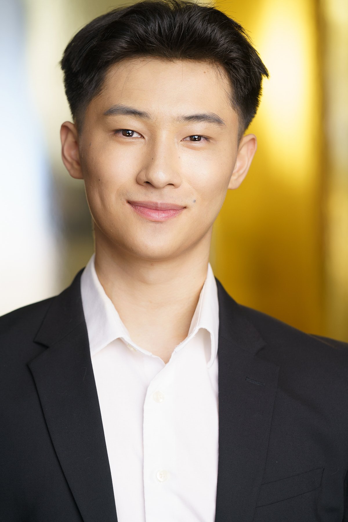 Young man in a black suit with a white shirt, smiling, against a blurred background with yellow and white colors.