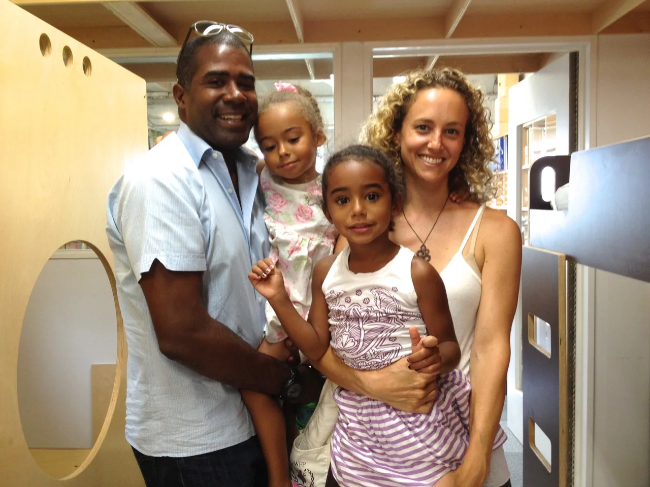 Four people indoors, faces obscured. Two adults on sides, two kids between. Background includes mirror and door/window.