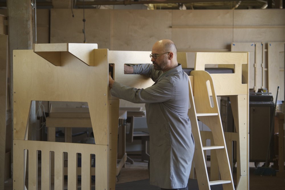 Man in a workshop inspecting wooden furniture parts, with tools and unfinished products around.