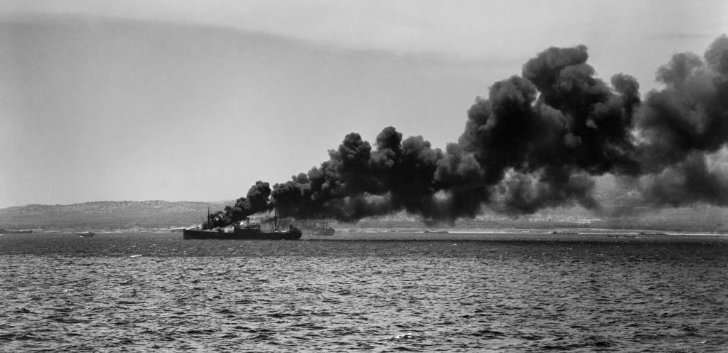 Operation Husky, invasion of Sicily, 10 July 1943: four miles south of Syracuse in 'Acid North' sector, an ammunition supply ship hit by enemy bombs burns close to the assault beaches. Landing craft can be seen on the shoreline below the smoke. (Lt F.G. Roper, R.N., Admiralty Official Collection, IWM A 18091).