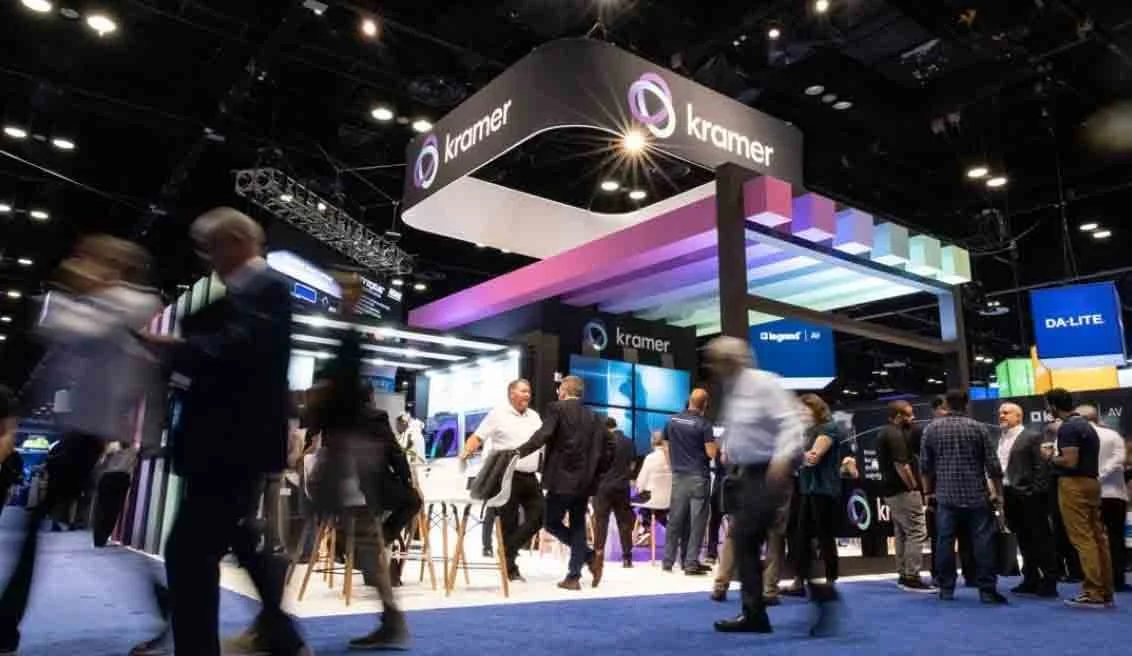 Trade show booth with the company name 'Kramer' and a colorful overhead structure, with people talking and walking around inside a large exhibition hall.