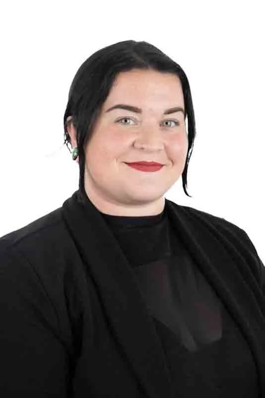 A woman with dark hair, fair skin, and red lipstick smiling, wearing a black top and blazer against a white background.