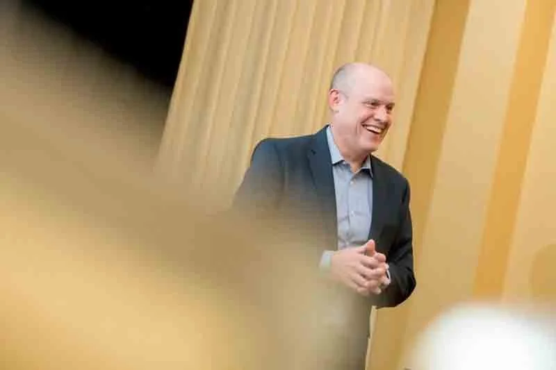 A man in a suit smiling and clapping in a room with wooden paneling walls.