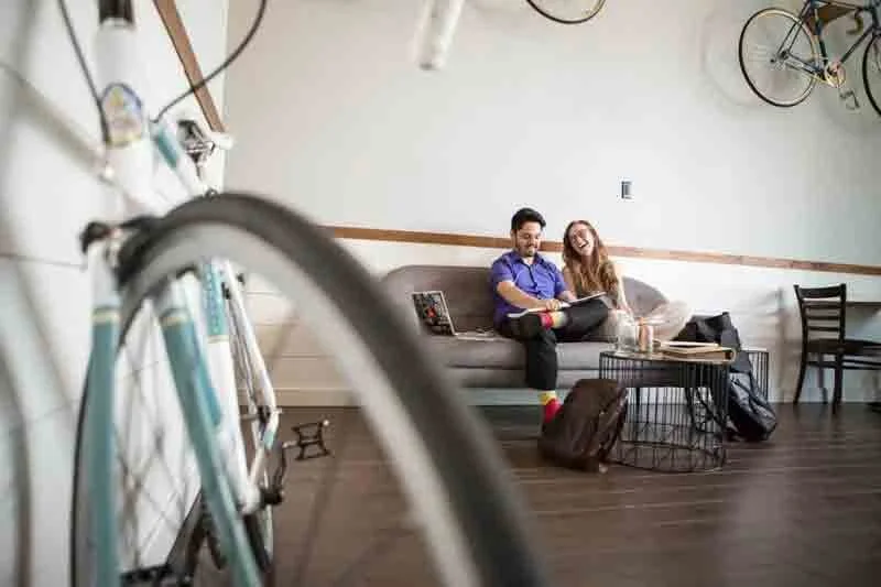 Two people sitting on a gray couch in a modern living room, smiling and looking at a laptop. A bicycle is partially visible in the foreground and hanging on the wall behind them is another bicycle. The room has a wooden floor and minimal decor.