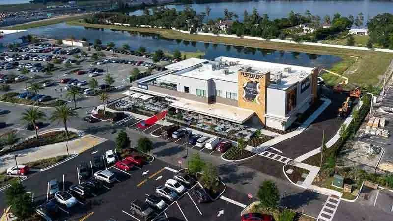 A large white commercial building with a sign that reads "Firestone" and an outdoor seating area, surrounded by parking lots filled with cars, near a body of water with green landscape in the background.