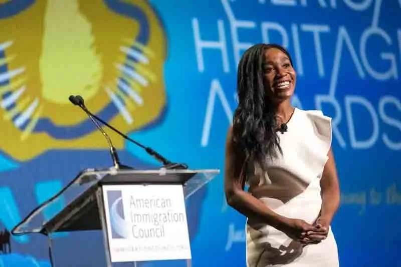 A woman smiling and standing on stage at the American Immigration Council awards event, with a podium in front of her that has a sign reading "American Immigration Council." The background features a large graphic with the words "Heritage Awards."
