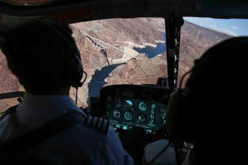 View from inside a helicopter cockpit showing two pilots flying over a dam and a reservoir in rugged terrain.