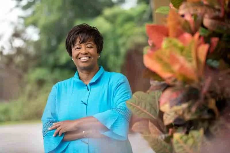 A woman wearing a bright blue shirt, smiling with arms crossed, standing outdoors with greenery in the background.
