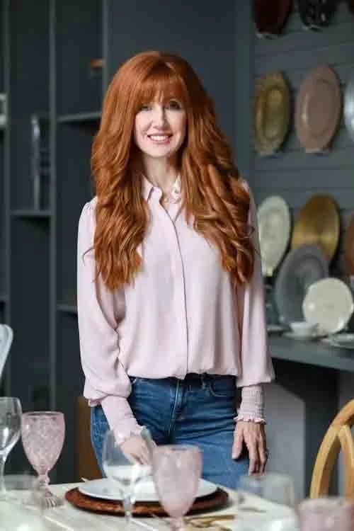 A woman with long, wavy red hair, wearing a light pink blouse and blue jeans, standing in a dining room with a table set with plates, glasses, and pink and white dishes on a sideboard in the background.