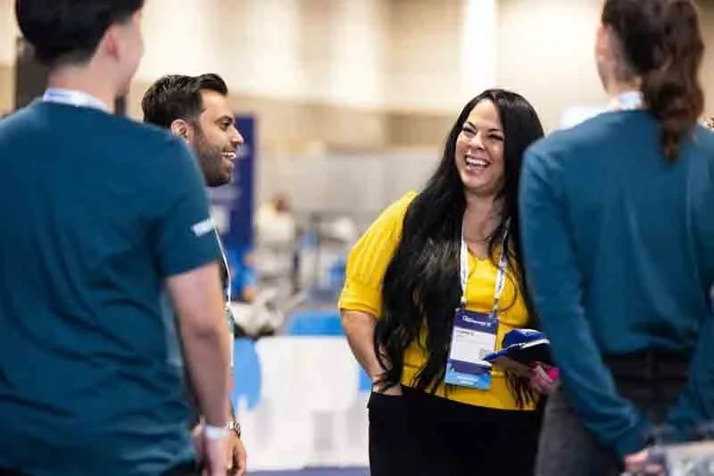 Four people, three women and one man, are smiling and talking at an indoor event or conference, with three wearing blue outfits and one woman in a yellow top.